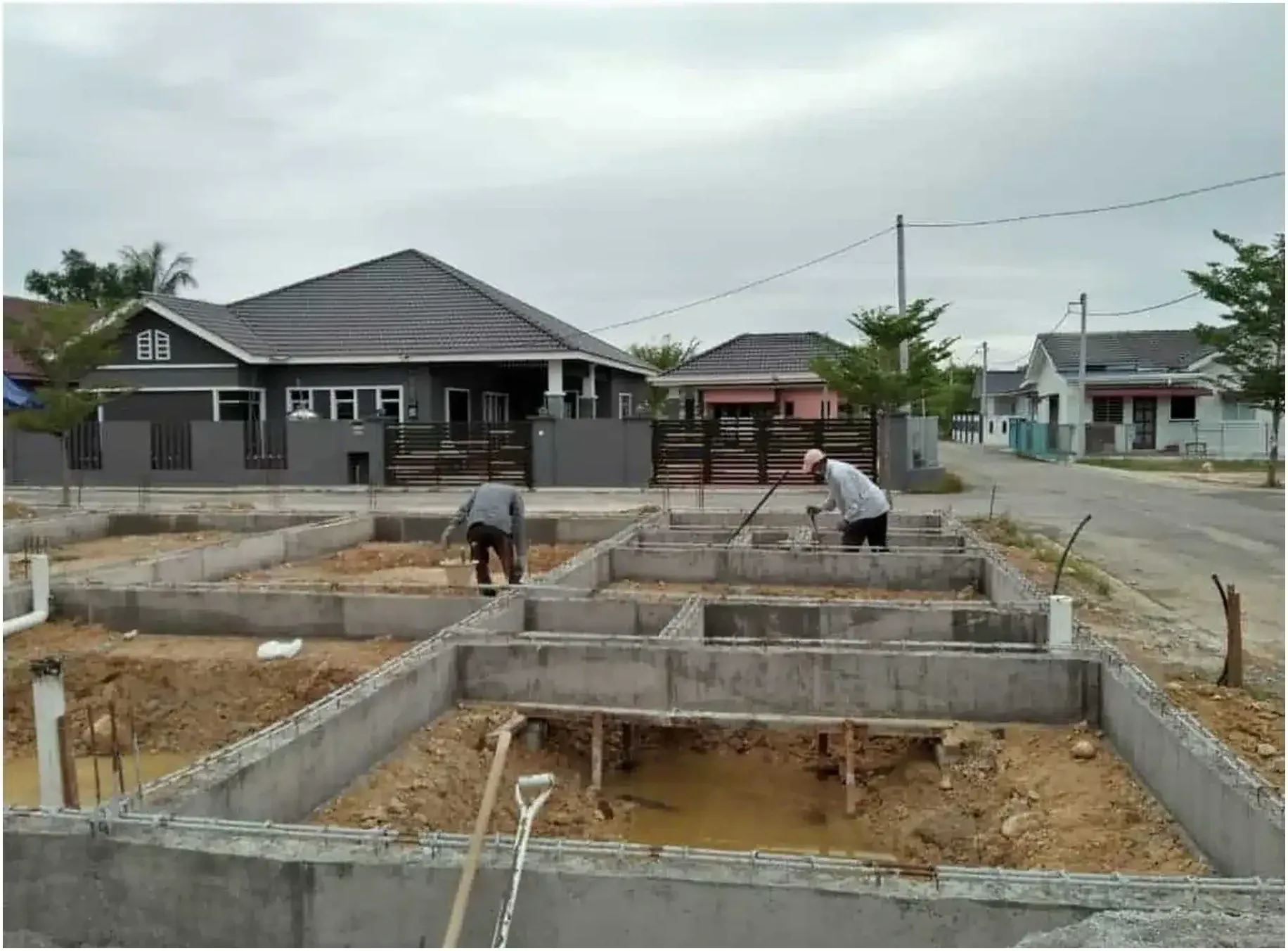 Site work on a RumahHQ house at foundation stage — workers levelling soil between finished concrete ground beams, on a residential street