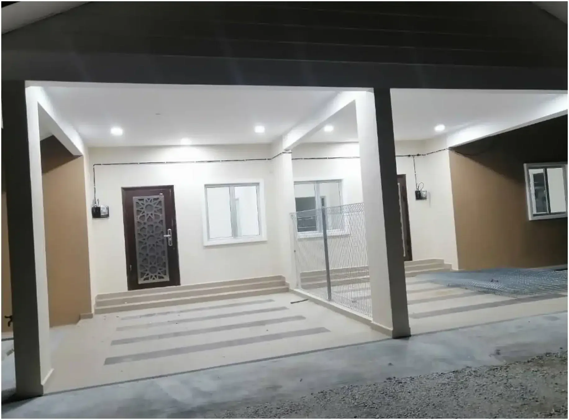 A finished RumahHQ porch at night — two meter boxes mounted on the wall with surface conduit, a steel grille door, and ceiling downlights already lit