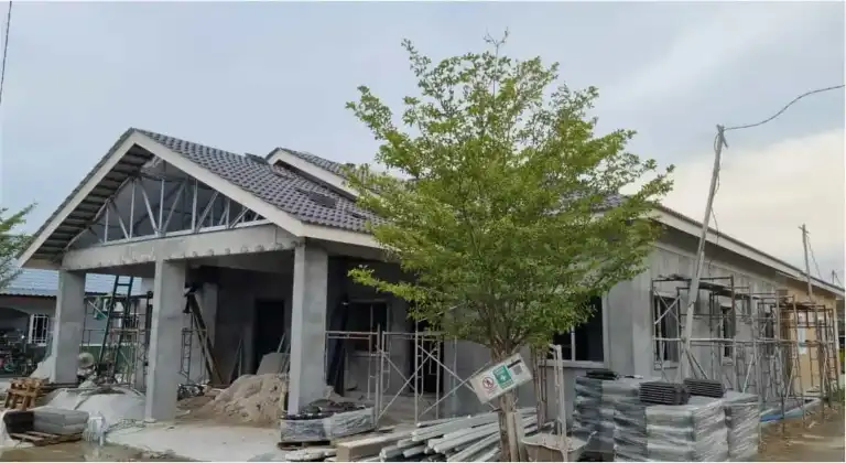 Rumah banglo RumahHQ pada peringkat struktur — kekuda keluli gable kelihatan di bahagian hadapan, genting mula dipasang di bahagian lain, tiang dan rasuk konkrit masih tanpa kemasan, dikelilingi perancah dan timbunan bahan