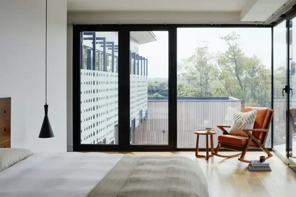 Modern bedroom with large windows, cozy chair, and wooden floors overlooking a lush view. Minimalist design.