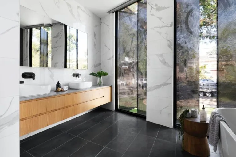 Modern bathroom with dual sinks, marble tiles, large mirror, and natural light through floor-to-ceiling windows.
