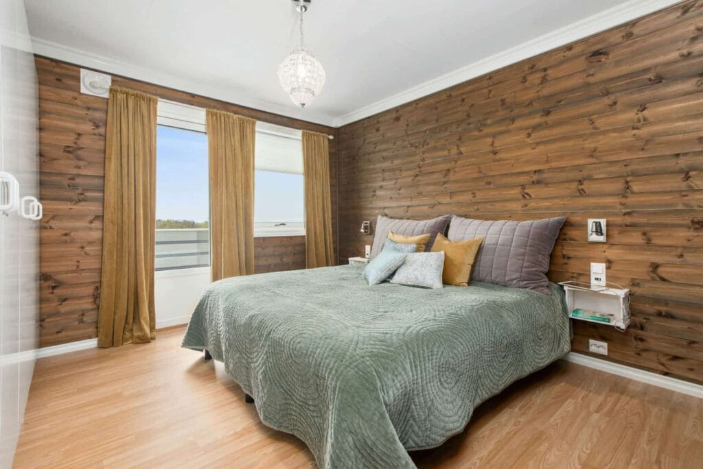 Cozy bedroom with wood panel walls, green bedspread, decorative pillows, and large window with brown curtains.