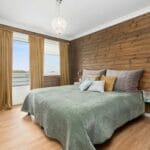 Cozy bedroom with wooden walls, green bedding, decorative pillows, and large window view.