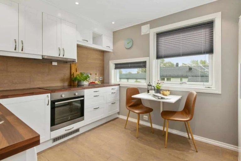Modern kitchen with white cabinets, wood countertops, and dining nook by a window. Brown chairs complete the cozy space.