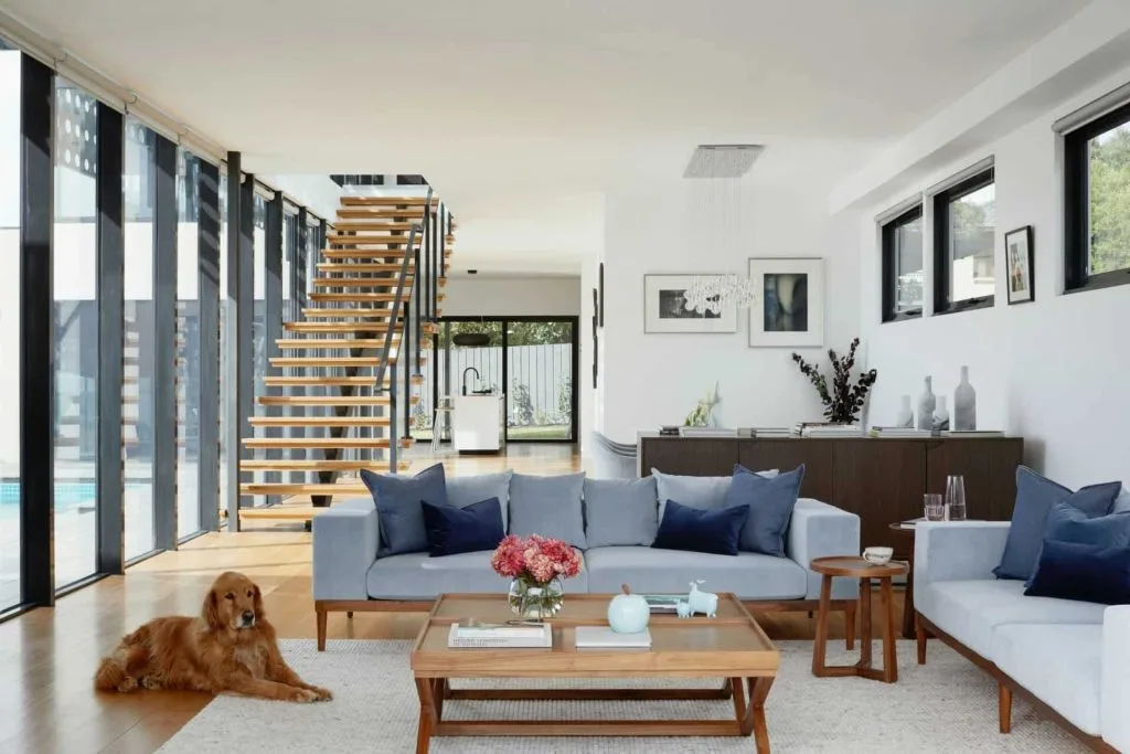 Modern living room with gray couches, wooden table, and a dog, featuring open staircase and floor-to-ceiling windows.