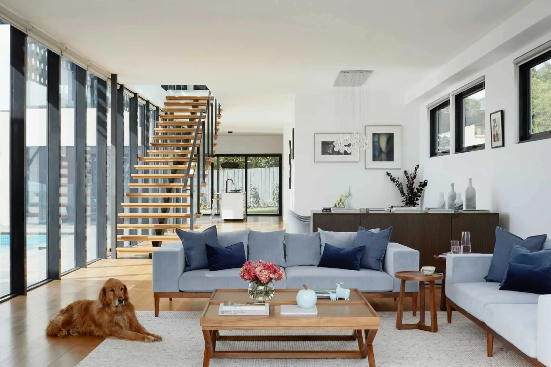 Modern living room with gray couches, wooden table, and a dog, featuring open staircase and floor-to-ceiling windows.
