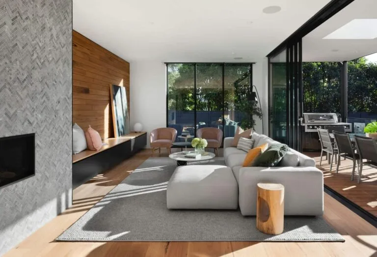 Modern living room with gray sofa, wood accents, and open patio doors leading to outdoor dining area.